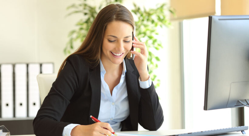 Happy businesswoman calling on mobile phone and taking notes on a desk at office